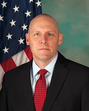 Portrait of Mr. Leland Kent Shea wearing a black suit with red tie and white shirt, set on a background of light blue with the United States flag running through the left half.